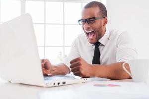 trabajo demasiado estresante. furioso joven africano con camisa y corbata gritando mientras mira la laptop mientras se sienta en su lugar de trabajo foto