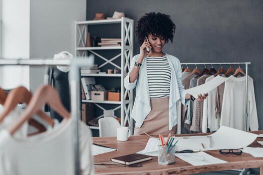 siempre disponible. hermosa joven africana hablando por teléfono inteligente y sonriendo mientras está de pie en su taller foto