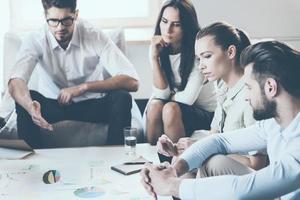 Finding solution together. Group of young business people discussing something while sitting together and looking at paper laying on desk photo