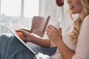 Perfect way to find fresh news. Close up of young woman using digital tablet and smiling while sitting on the sofa at home photo