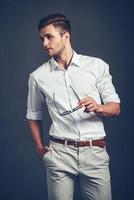 Confident in his style. Confident young handsome man carrying his sunglasses and looking away while standing against grey background photo