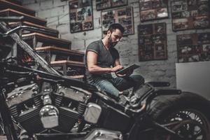 planeando su semana. joven confiado sosteniendo una tableta digital y mirándola mientras se sienta cerca de la motocicleta en el taller de reparación foto