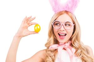 este huevo de pascua es perfecto, hermosa mujer joven de cabello rubio con orejas de conejo sosteniendo un huevo de pascua y mirándolo con una sonrisa mientras está aislada en el fondo blanco foto