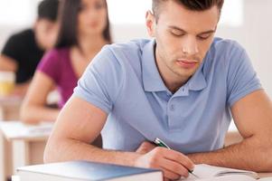 estudiante confiado. guapo estudiante masculino escribiendo algo en el bloc de notas mientras está sentado en el aula con otros estudiantes foto