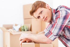 Tired of moving. Side view of depressed young man leaning at the cardboard box and expressing negativity while more boxes laying in the background photo