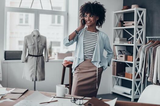 buena charla con el cliente. atractiva joven africana hablando por teléfono inteligente y mirando hacia otro lado con una sonrisa mientras estaba en su taller foto