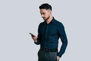 Sending important business messages. Serious young man in shirt typing text message using his smart phone while standing against grey background photo