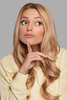 Trying to find an answer Confused young woman keeping index finger on her face while standing against grey background photo