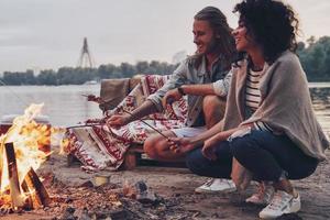 No cares. Group of young people in casual wear roasting marshmallows over a campfire while resting near the lake photo