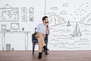 Your dreams can send you far away. Young handsome man keeping hand on chin and looking away while sitting in the chair against illustration of fjord vs. working place photo