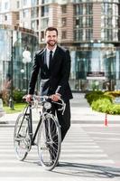 la manera perfecta de llegar al trabajo. longitud total de un joven hombre de negocios sonriente rodando su bicicleta mientras cruza la calle foto