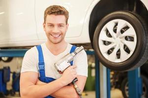Ready to work. Confident young man holding a pneumatic wrench and smiling while standing in workshop with car in the background photo