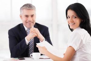 Woman on job interview. Cheerful young woman looking over shoulder and smiling while mature man in formalwear sitting on background photo