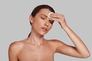 Feels good... Attractive young woman applying cotton pad and keeping eyes closed while standing against grey background photo