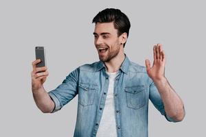 Hello Good looking young man in blue jeans shirt waving and smiling while looking at his smart phone and standing against grey background photo