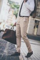 Trendy look. Part of man holding leather bag while walking outdoors photo