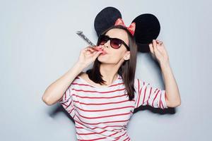 She clearly loves holidays  Playful young woman in glasses and mouse ears blowing in noisemaker while standing against grey background photo