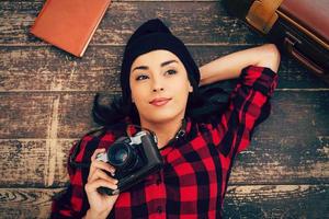 Beautiful day dreamer. Top view of beautiful young woman in headwear lying on the floor and holding camera while suitcase and note pad laying near her photo