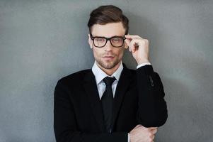 Stylish handsome. Confident young businessman adjusting his eyewear and looking at camera while standing against grey background photo