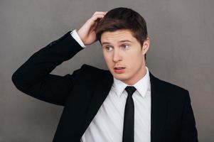 He is not sure about it. Frustrated young man in formalwear holding hand in hair and looking away while standing against grey background photo