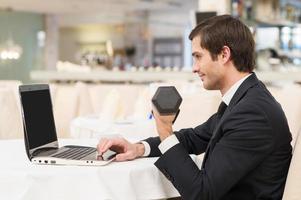 Sporty businessman. Cheerful young man in formalwear sitting at laptop and holding a dumbbell photo