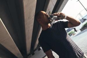 Hydrating. Young African man in sports clothing drinking water while standing outside photo