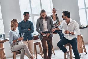 feliz de ser un equipo. grupo de jóvenes modernos con ropa informal inteligente sonriendo y discutiendo algo mientras trabajan en la oficina creativa foto