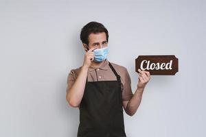 Displeased young man in apron wearing protective face mask and holding close sign while standing against gray background photo