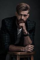 Stylishly confidence. Handsome young man in casual clothing looking away while leaning on stool against grey background photo