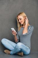 Great message from friend. Beautiful young woman using her smart phone and looking at it with smile while sitting in lotus position against grey background photo