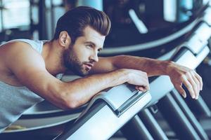 After five miles. Side view of young man in sportswear looking exhausted while leaning on treadmill at gym photo