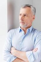 Thinking about solutions. Thoughtful grey hair senior man in shirt looking away while standing near the window photo