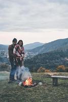 Fire keeps them warm. Handsome young man embracing his attractive girlfriend while standing near the campfire outdoors photo