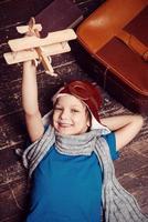 Little day dreamer. Top view of happy little boy in pilot headwear and eyeglasses lying on the hardwood floor and playing with wooden planer photo