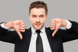 Bad news for you. Confident young man in formalwear showing his thumbs down and looking at camera while standing against grey background photo