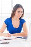 So much work to do. Confident young woman writing something at her note pad and looking at laptop while sitting at her working place photo