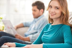 Couple at home. Beautiful young woman sitting on the couch and smiling at camera while her husband sitting on the background and reading newspaper photo