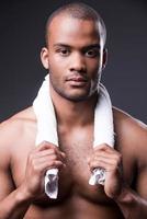 Relaxing after training. Young shirtless African man carrying towel on shoulders and looking at camera while standing against grey background photo