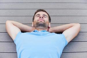 tiempo para relajarse. vista superior de un joven apuesto que mantiene los ojos cerrados y sonríe mientras se acuesta en el suelo de madera dura foto