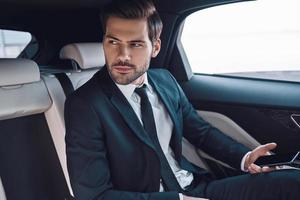 Young and successful. Handsome young man in full suit looking away while sitting in the car photo