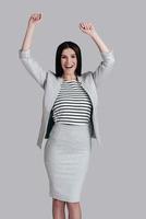 She is a winner Happy attractive young woman in smart casual wear keeping arms raised and looking at camera while standing against grey background photo