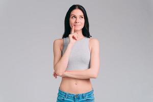 Thoughtful beauty. Thoughtful young woman holding hand on chin and looking away with toothless smile while standing against grey background photo