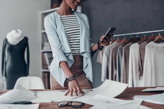buenos comentarios del cliente. primer plano de una joven africana sosteniendo un teléfono inteligente y sonriendo mientras estaba de pie en su estudio cerca de la ropa colgada en los estantes foto