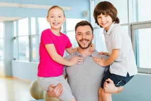 padre deportivo con hijos. alegre joven padre deportivo llevando a sus hijos en las manos y sonriendo mientras está de pie en el club deportivo foto