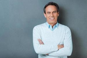 Confident mature man. Confident mature man looking at camera and smiling while keeping arms crossed and standing against grey background photo