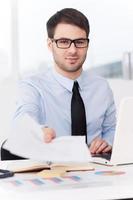 I need your signature her. Confident young man in shirt and tie stretching out documents and looking at camera while sitting at his working place photo