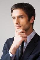 Thinking about solutions. Handsome young businessman looking away and holding hand on chin while standing against grey background photo