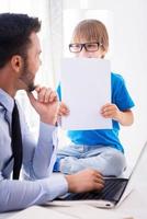 Feeling playful. Cheerful young man in shirt and tie working on laptop and looking at his son sitting close to him and holding documents photo