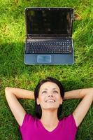 Living in digital age. Top view of beautiful young woman lying in grass and smiling while while laptop laying upon her head photo