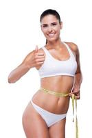 Her personal achievement. Attractive young woman in white bra and panties showing her thumb up while measuring her body with tape and isolated on white photo